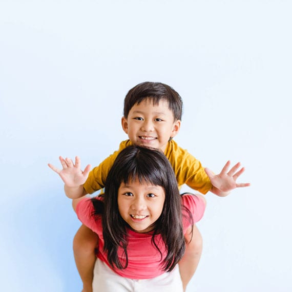 A young child in a yellow shirt sits on the shoulders of an adult in a pink top against a plain light blue background.