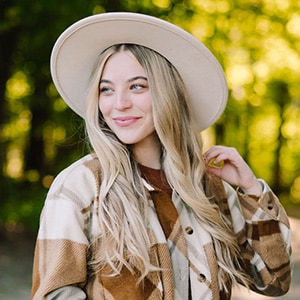 Kalynn in a white hat and checkered jacket touches her hair, standing outdoors with blurred green trees in the background.