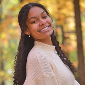 Katelyn wearing a cream sweater stands in a forest with autumn-colored leaves.